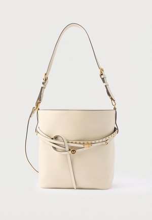 Geantă tip bucket din piele bej, cu curea reglabilă, capse aurii și detaliu decorativ cu curea în față, pe un fundal simplu.