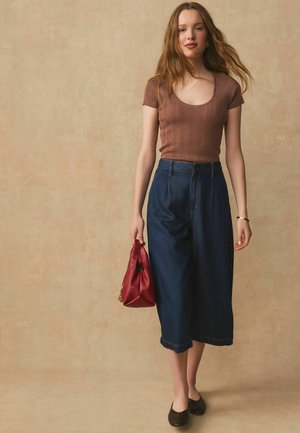 Tânără purtând un top maro din tricot cu mâneci scurte, culotte din denim albastru închis cu picioare largi, balerini negri, ținând o poșetă roșie mică pe un fundal bej.