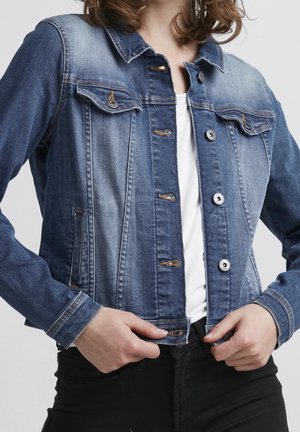 Femme portant une veste en jean bleue par-dessus une chemise blanche, en train de boutonner le bouton du bas, avec un pantalon noir visible.