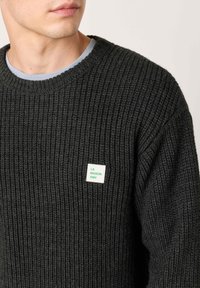 Tamnozeleni rebrasti džemper s okruglim izrezom i svijetloplavim unutarnjim ovratnikom, s bijelom etikietom na kojoj piše "LA MAISON AMV."