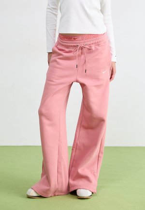 Persoon draagt roze wijde joggingbroek met trekkoord en witte sneakers, staand op een groene vloer tegen een witte muur.