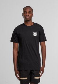 Svart bomull t-skjorte med korte ermer og rund hals, med et sirkulært brodert motiv på venstre bryst som viser en katt.