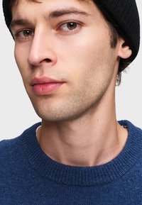 Modello che indossa un maglione lavorato a maglia blu con collo a girocollo a coste, in primo piano con focus sulle caratteristiche del volto e sulla texture del maglione.