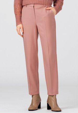 Vrouw die hoge, recht gesneden broek in stoffig roze, een malvekleurige gebreide trui en beige enkellaarsjes met hak draagt, staand tegen een lichte achtergrond.