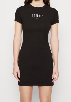 Kobieta w dopasowanej czarnej sukience z krótkim rękawem z napisem "Tommy Jeans" i logo na piersi, stojąca na białym tle.