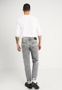 Man med tatuering på vänster arm bär en vit långärmad skjorta, grå slitna jeans, vita strumpor och svarta skor, står med ryggen vänd mot kameran.