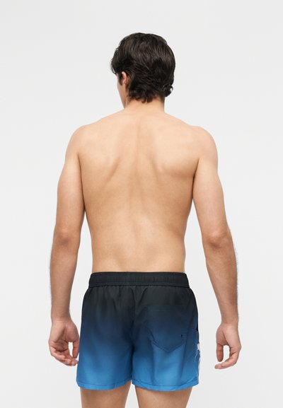 Homme portant un short de bain à dégradé bleu et noir, debout torse nu, dos tourné vers la caméra sur un fond clair uni.