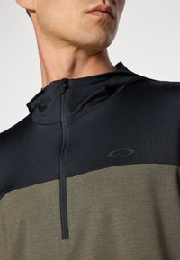 Quarter-zip hettegenser i svart og olivengrønn. Har en glatt tekstur, kontrasterende farger, og en diskret logo på brystet.