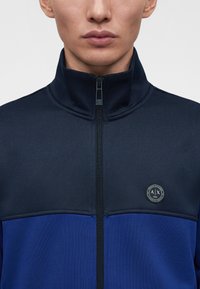 Мъж, носещ тъмносиньо и синьо яке с цип и висок яка, с кръг логото A|X Armani Exchange на гърдите.