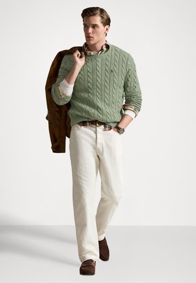 Green cable knit sweater over a plaid shirt, white pants, and brown suede shoes. Model holds a brown jacket and wears a watch.