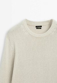 Pullover lavorato a maglia beige chiaro, con scollo rotondo, polsini a costine e finitura texturizzata, con un'etichetta visibile all'interno del colletto.