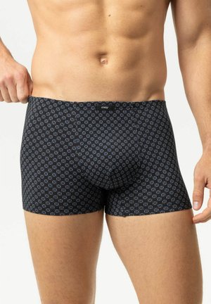 Männlicher Oberkörper trägt schwarze Boxershorts mit blauem geometrischem Quadrat-Muster und zieht den Bund an einer Seite leicht nach außen.