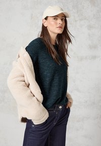 Dunkelblaues Strickoberteil unter einem cremefarbenen, künstlichen Schaffellmantel. Dunkle Jeans mit Knopfdetail. Inklusive einer hellen Mütze.