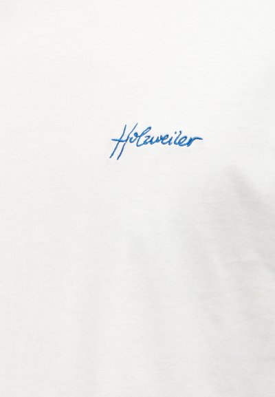 Λευκό ύφασμα με μικρό μπλε κεντημένο κειμενόγραφο "Holzweiler" στο κέντρο της επιφάνειας.