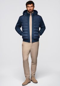 Giubbotto imbottito blu navy con cappuccio e polsini a coste, indossato sopra un maglione beige e pantaloni color beige, abbinato a stivali alla caviglia marroni.