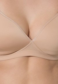 Soutien-gorge sans fil beige avec une finition lisse, des bonnets triangulaires et un éclat subtil. Présente des accents décoratifs près du centre et des bandes douces et flexibles.