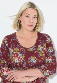 Bordeauxfarbene Bluse mit floralem Muster und kurzen Ärmeln, die rote und blaue Blumen zeigt, mit einem runden Ausschnitt und einer weichen, baumwollähnlichen Textur.