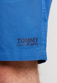Modré šortky z hladkého materiálu mají viditelné logo "TOMMY JEANS" na dolním levém lemu, s kontrastním žlutým šitím.