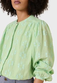 Pale green blouse with puffed sleeves, embroidered floral patterns in matching thread, and a button closure at the neck. Textured fabric.