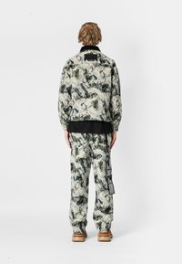 Chaqueta con capucha y pantalones a juego en una tela texturizada con un patrón abstracto en verde y beige. Presenta un acento negro en el cuello.