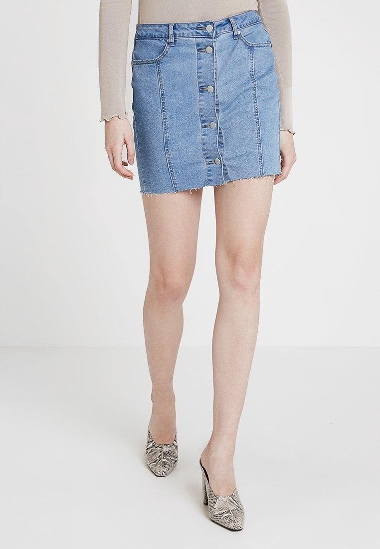 Mini-jupe en denim bleu clair avec un ourlet brut, dotée d'une fermeture à boutons sur le devant et de deux poches avant. Portée avec des chaussures à talons à motifs.