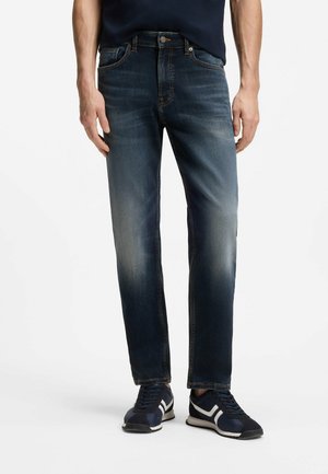 Homme portant un jean bleu foncé délavé et des baskets bleu marine avec des rayures blanches, debout devant un fond blanc uni.