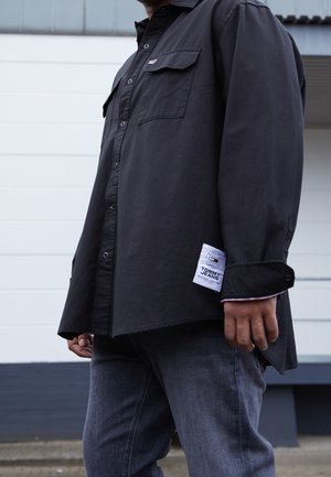 Persona che indossa una camicia nera a maniche lunghe con bottoni e toppa Tommy Jeans, abbinata a jeans in denim grigio, in piedi all'aperto.