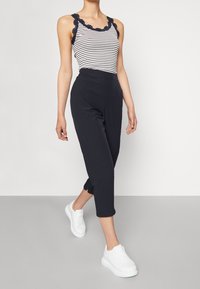 Pruhovaný top na ramínka s námořnickými volány, doplněný tmavými zkrácenými kalhotami a bílými teniskami. Hladké textury, ležérní design.