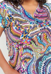 Blusa a maniche corte con zip, caratterizzata da un vivace motivo a fiori astratti e geometrici multicolori in blu, rosa, giallo e verde.