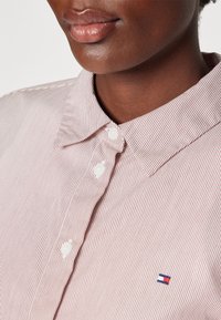 Zbliżenie osoby noszącej jasnoróżową koszulę na guziki w białe paski z małym czerwono-biało-niebieskim logo flagi na piersi.