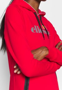 Personne portant un sweat à capuche ellesse rouge avec des cordons noirs, bras croisés, sur un fond blanc uni.