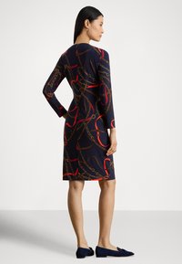 Žena stojaca v profile, oblečená v tmavomodrých šatách s dlhým rukávom, ktoré majú červené a hnedé vzory reťazí a lán, v kombinácii s tmavomodrými balerínovými topánkami.