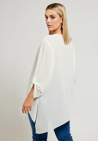 Vit blus med långa ärmar och hög-låg fåll, med rullade ärmar och en mjuk, lättviktig tyg. Sidoklipp för ökad rörelsefrihet.
