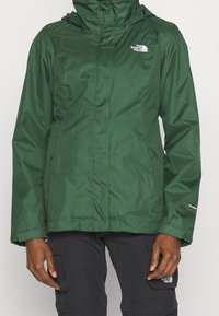 Giacca impermeabile verde scuro con zip frontale, cappuccio regolabile, tasche laterali e logo sul petto. Texture liscia e design aderente.
