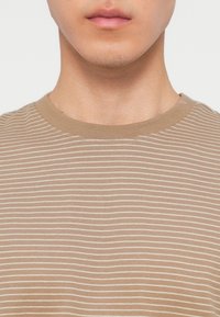 Κοντινό πλάνο του κάτω μέρους του προσώπου και του λαιμού ενός ατόμου που φοράει μπλούζα με στρογγυλή λαιμόκοψη σε μπεζ και λευκές ρίγες.