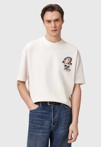 Junger Mann trägt ein weißes übergroßes T-Shirt mit einem bunten Drachen- und Totenkopf-Print auf der linken Brust, kombiniert mit blauen Jeans und einem schwarzen Gürtel.