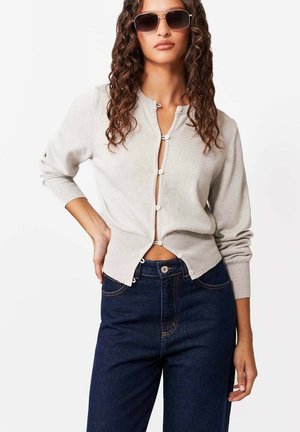 Kobieta o kręconych włosach, ubrana w beżowy kardigan zapinany na guziki, ciemnoniebieskie jeansy z wysokim stanem oraz duże ciemne okulary przeciwsłoneczne, pozująca z jedną ręką na biodrze.