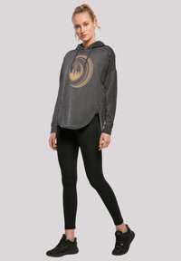 Fekete kapucnis pulóver kerek szegéllyel és arany kör alakú grafikai designnal. Fekete leggings-szel és fekete sportcipőkkel párosítva.