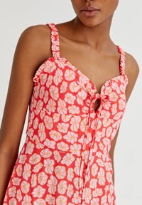 Frau trägt ein ärmelloses Kleid mit rosa und orangefarbener Blumenmusterung, das über Rüschen-Träger und einen vorderen Knoten verfügt.