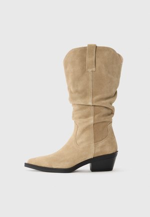 Beige suède cowboylaars met een slouchy schacht, puntige neus en blokhak. Vo smooth textuur met minimale stiksels en een detail met een trekband aan de achterkant.