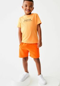 Orange kortærmet t-shirt med sort "BAKER" tekst, parret med klare orange shorts. Hvide sneakers fuldender outfittet. Glat stoftekstur.