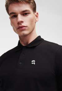 Polo shirt nera in tessuto testurizzato, con colletto e tre bottoni. Un piccolo logo bianco è ricamato sul lato sinistro del petto.