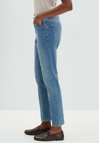 Vaaleansiniset suorat farkut, jotka on valmistettu denimestä, ja joissa on viisi taskua sekä vähäistä kulutusta, yhdistettynä ruskeisiin liukuselkimiin.