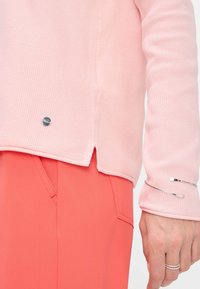 Helles rosa strukturiertes Sweatshirt mit einem seitlichen Schlitz, versehen mit einem kleinen markierten Metallakzent am Saum, kombiniert mit korallenfarbenen Hosen und Silber-Schmuck.