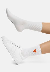 Witte geribbelde crew-sokken met een oranje hartontwerp, gecombineerd met witte sneakers met een getextureerde zijpaneel en vetersluiting.