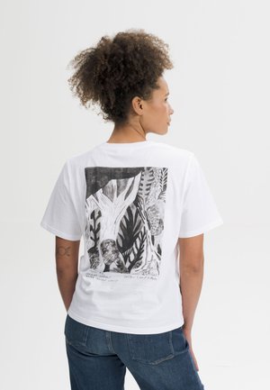 Vrouw met krullend haar die een wit T-shirt draagt met een zwart-wit natuurthema op de rug en een blauwe spijkerbroek.
