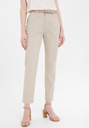 Femme portant un pantalon beige droit avec une ceinture tressée et des sandales à talons blocs marron clair, debout sur fond blanc.