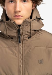 Giacca imbottita marrone con colletto alto, zip frontale, cordini e una toppa con logo sul petto. Tessuto liscio e design su misura.