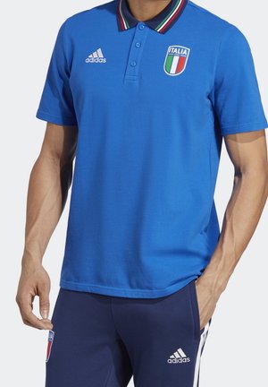 Mężczyzna ubrany w niebieską koszulkę polo Adidas z logo narodowej reprezentacji Włoch w piłce nożnej oraz dopasowane niebieskie spodnie Adidas z białymi paskami i logo.