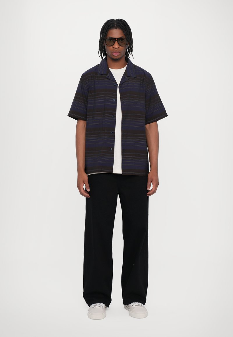 Chemise à manches courtes à rayures bleu foncé et noires, avec un avant boutonné, associée à un pantalon large noir et des baskets blanches. Des lunettes sont portées.